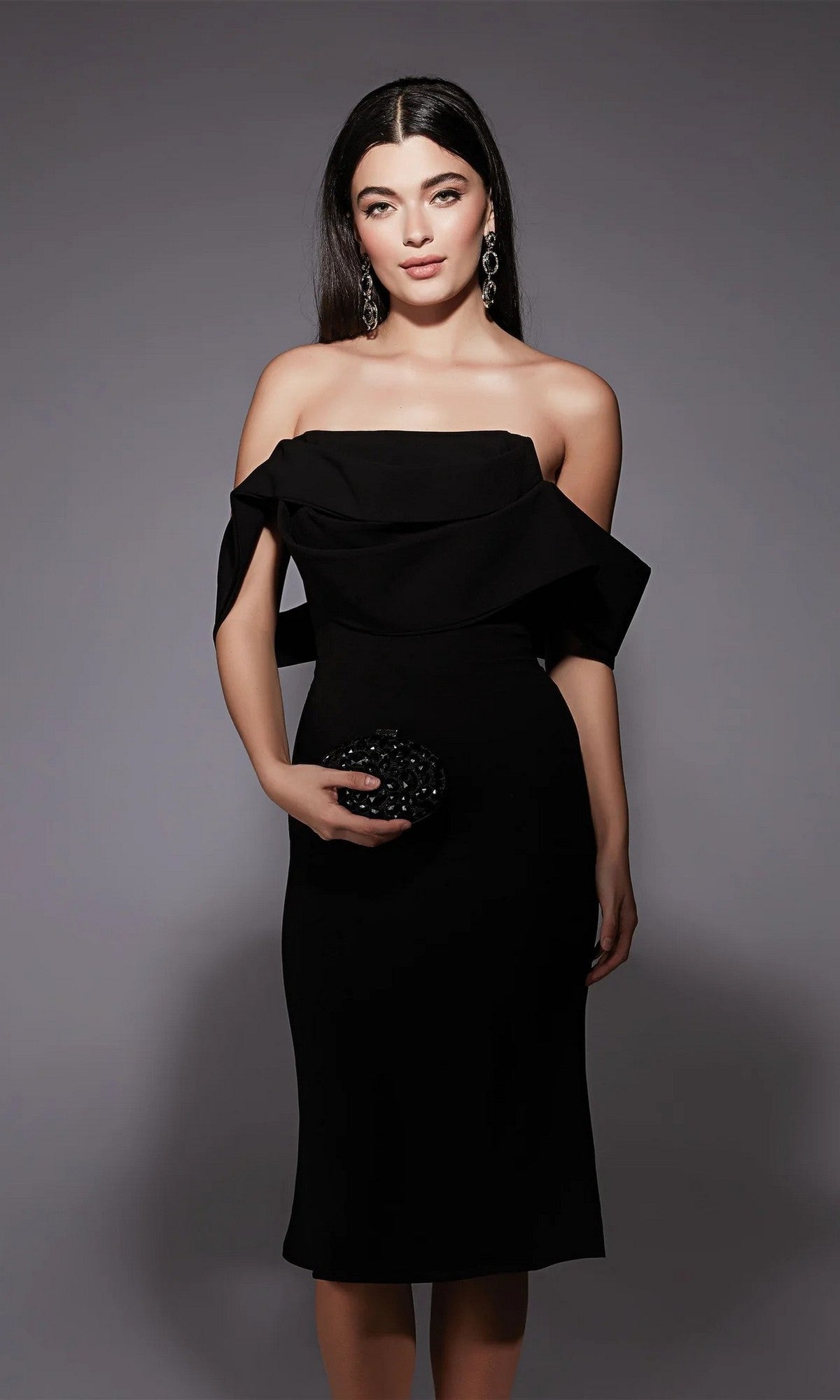 A woman in the black Alyce 70055 knee-length semi-formal dress and statement earrings poses against a gray background, holding a small black clutch.